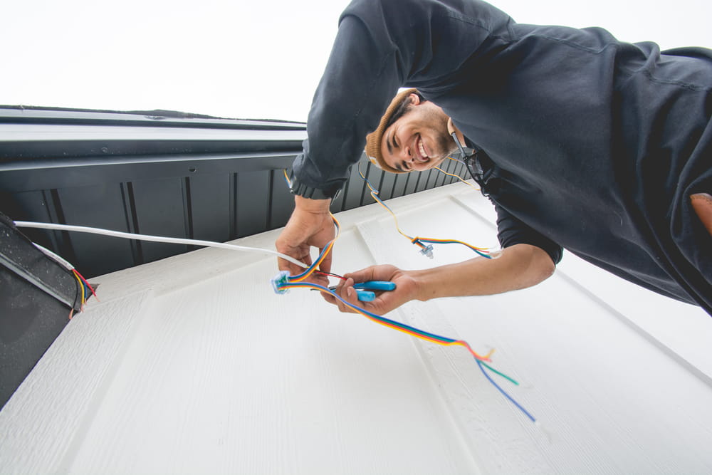 Attached image file A person smiling while installing or repairing electrical wiring on an exterior wall, holding colorful wires and using tools, viewed from a low angle.