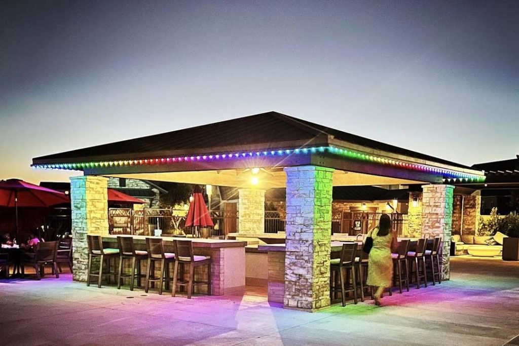 Attached image file An outdoor bar with stone pillars and a dark roof is lit by multicolored LED lights at dusk. Wooden barstools surround the bar, and a person walks nearby. Red umbrellas and seating are visible in the background.
