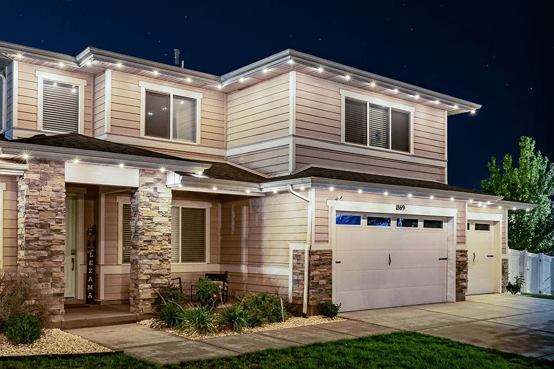 Attached image file Two-story suburban house at night with stone accents, beige siding, a double garage, and warm outdoor lighting outlining the roof. Small shrubs and plants line the front yard.