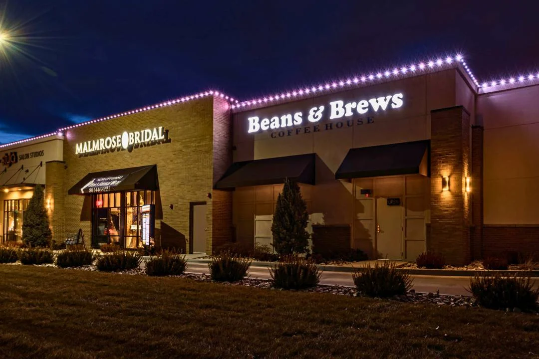 Attached image file A well-lit storefront at night features two businesses: Malmrose Bridal, with a black awning, and Beans & Brews Coffeehouse, both with illuminated signs. Low shrubs and grass line the walkway in front of the buildings.