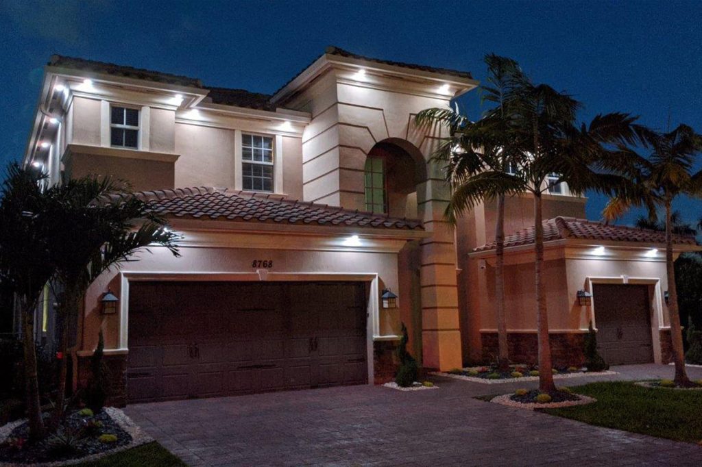 Attached image file A modern two-story house with exterior lights illuminating the beige walls and brown tiled roof at night, surrounded by palm trees and neatly landscaped greenery.