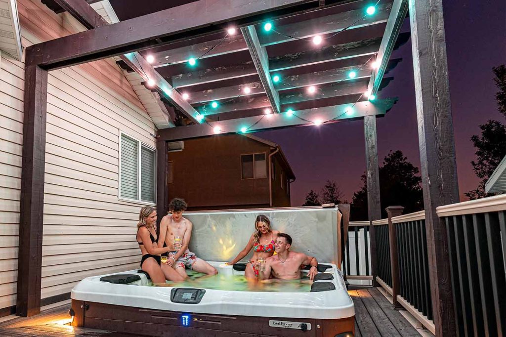 Attached image file Four people in swimsuits relax in a hot tub on a deck at dusk, under a pergola decorated with string lights. Two women and two men are enjoying the water, with a house and trees visible in the background.