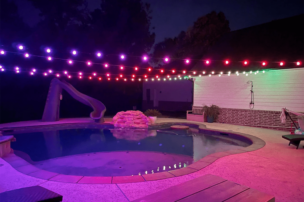 Attached image file A backyard pool at night is lit by strings of purple, red, and white lights overhead. A slide leads into the pool, and a small rock waterfall feature sits beside it. The area is quiet and empty.