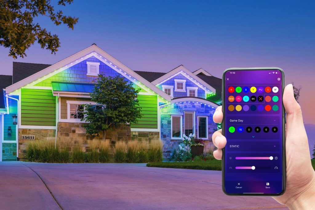 Attached image file A hand holds a smartphone displaying an app for controlling outdoor house lights, which are illuminated in colorful patterns on the exterior of a modern suburban home at dusk.