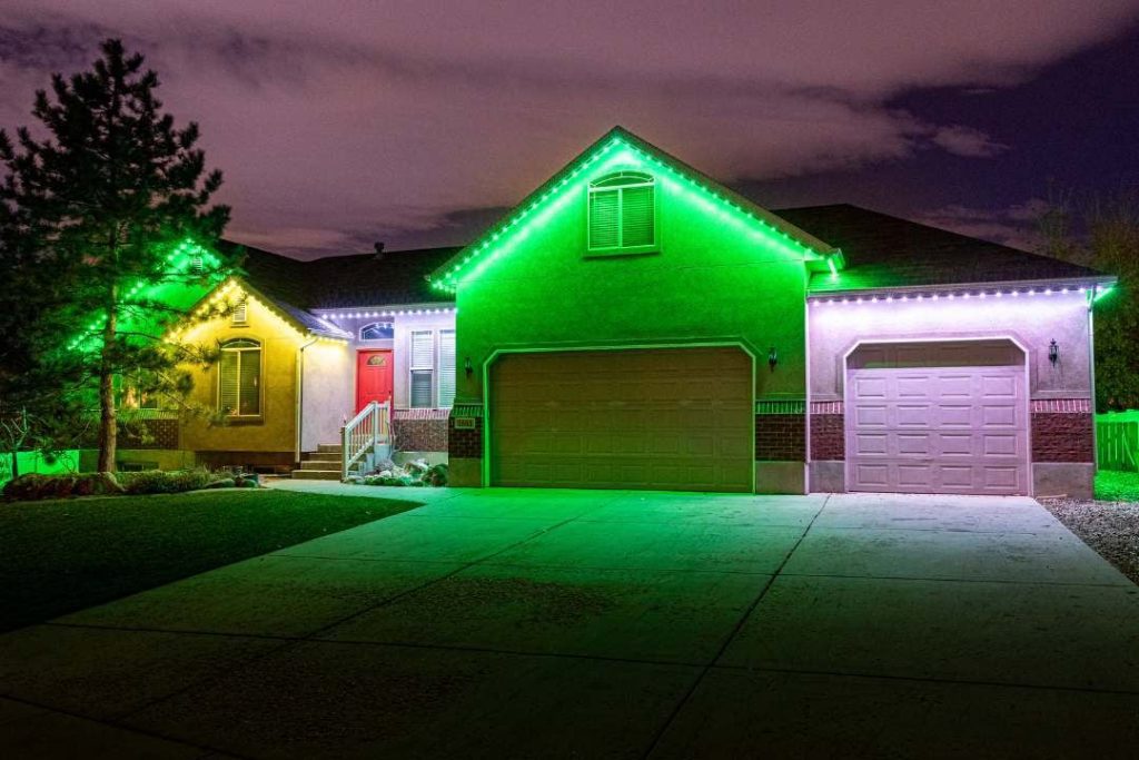 Attached image file A single-story house at night is lit with green and white LED lights along the roofline, highlighting the garage doors, entryway, and front yard. The sky is cloudy, and a tree stands on the left side of the house.
