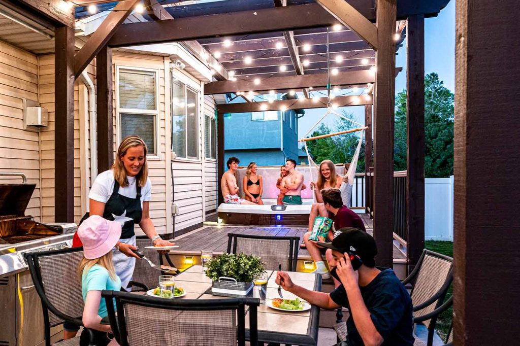 Attached image file A woman serves food at an outdoor patio table to two children, while four adults relax and chat in a hot tub nearby. String lights hang overhead, and the scene is cheerful and lively in a backyard setting.