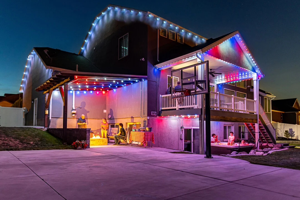 Attached image file A two-story house at dusk decorated with colorful string lights. People are gathered on the patio and under the balcony, enjoying an outdoor social event next to a basketball hoop.
