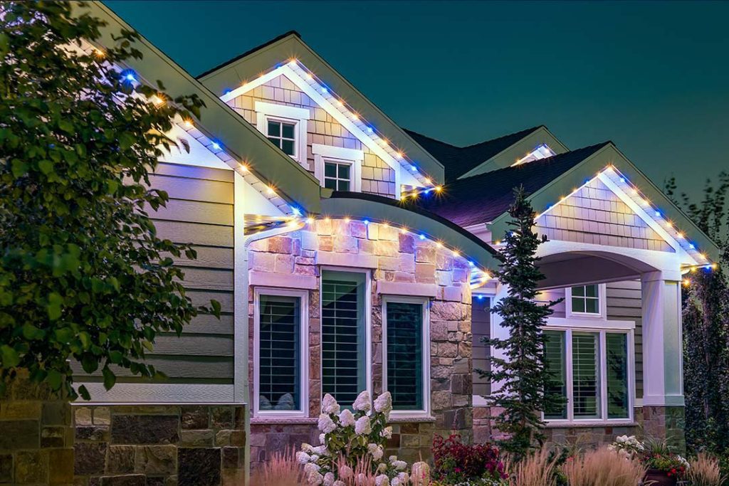 Attached image file A modern house at dusk with a stone and siding exterior, white-trimmed windows, landscaped garden, and blue and white string lights outlining the roof.