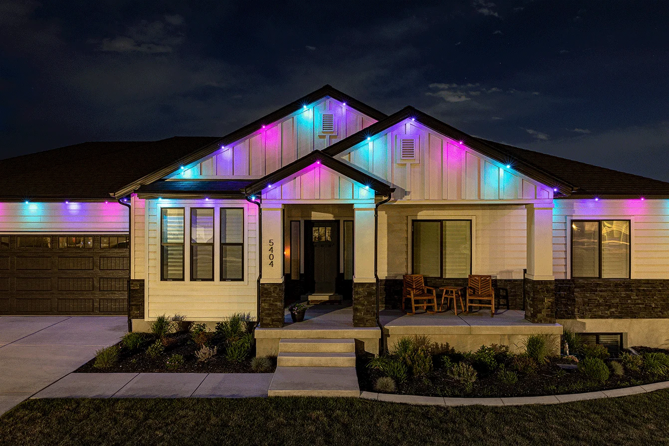 Attached image file A modern single-story house at night, decorated with colorful blue, pink, and purple lights along the roofline. The front porch has two wooden chairs and is surrounded by neatly trimmed bushes.