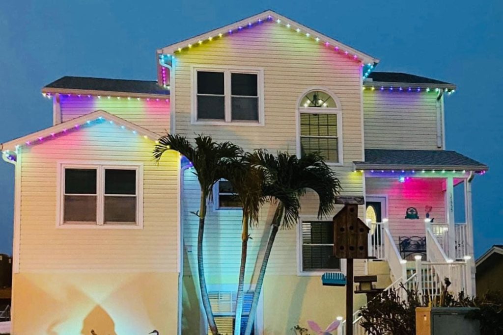 Attached image file A two-story house is decorated with colorful string lights along the roof edges, glowing in the evening. Two palm trees stand in front, and a birdhouse is visible near the staircase on the right.