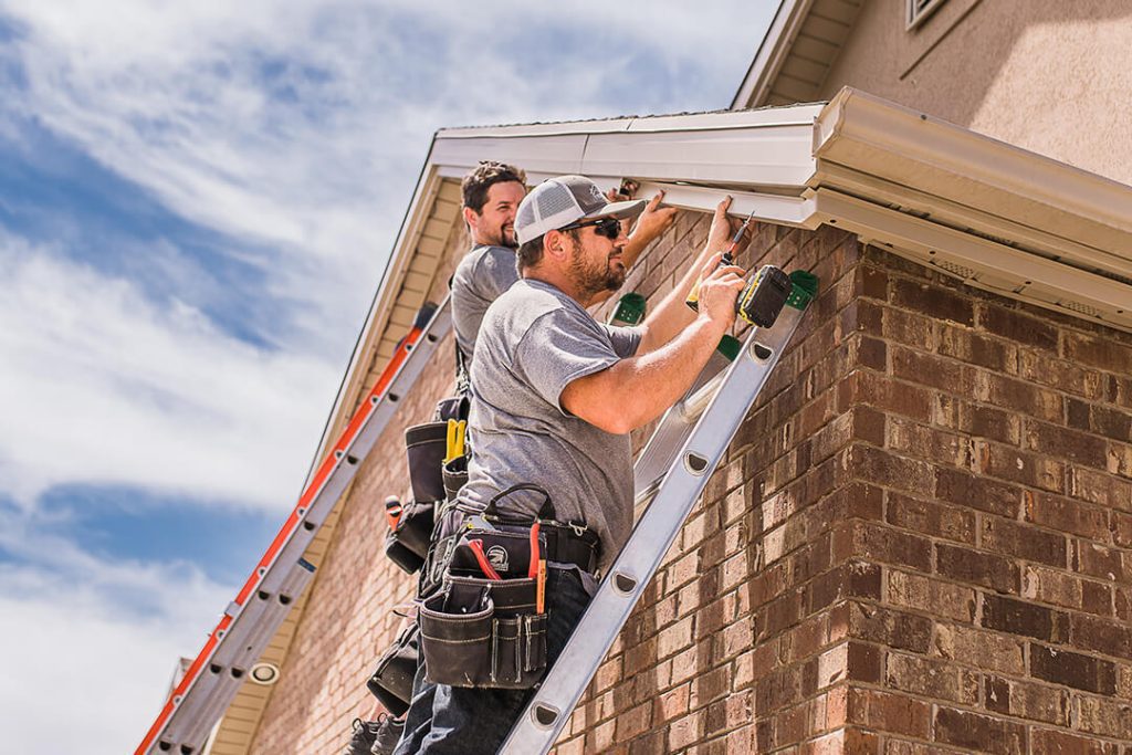 Attached image file Two workers wearing tool belts stand on ladders, installing or repairing gutters on the roof edge of a brick house under a partly cloudy sky.