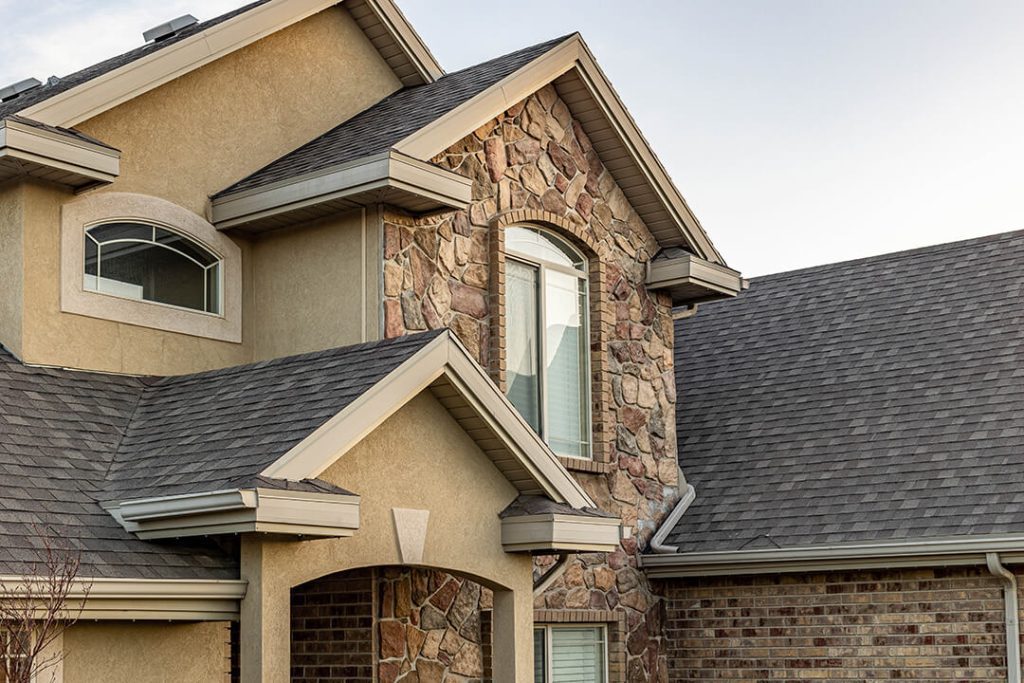 Attached image file A close-up of a suburban house exterior featuring a mix of stone, brick, and stucco siding with multiple gables and grey shingle roofing. Several windows are visible under a clear sky.