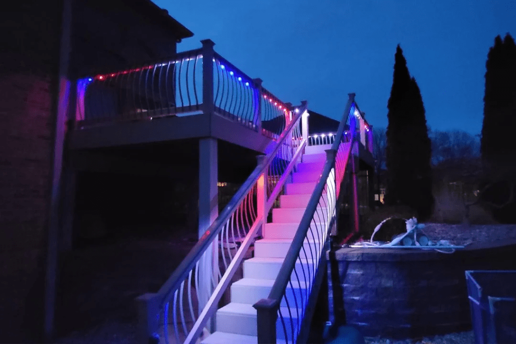 Attached image file A staircase leading to an elevated deck is illuminated by red, white, and blue lights at night, creating a vibrant glow on the railings and steps. Trees and a garden area are visible in the background.