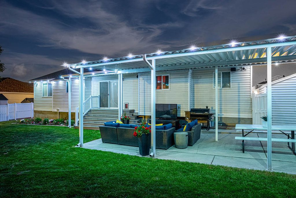 Attached image file A backyard patio at night with white overhead lights, outdoor seating, a grill, potted plants, and a picnic table on a concrete area beside a grassy lawn, behind a light-colored house.