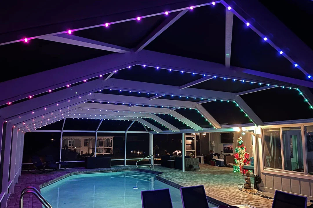 Attached image file A covered outdoor pool area at night is decorated with colorful LED strip lights along the roof. There is patio furniture around the pool and a lit Christmas tree near the back.