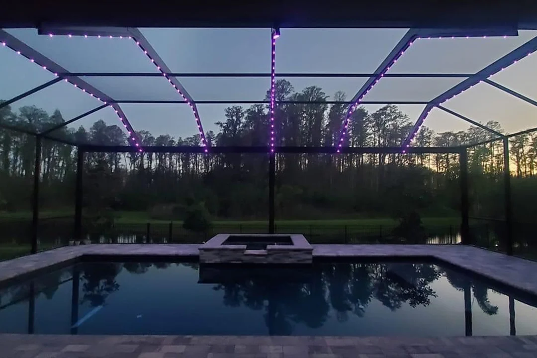 Attached image file A screened-in pool area at dusk, featuring purple string lights outlining the frame, with trees and their reflection visible in the water and a sunset sky in the background.