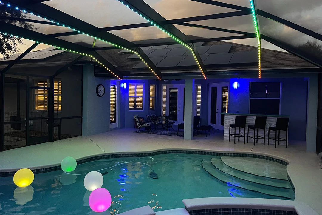 Attached image file A backyard pool at dusk features floating, glowing balls in the water. A covered patio with colorful LED lights, bar stools, and lounge seating is visible in the background.