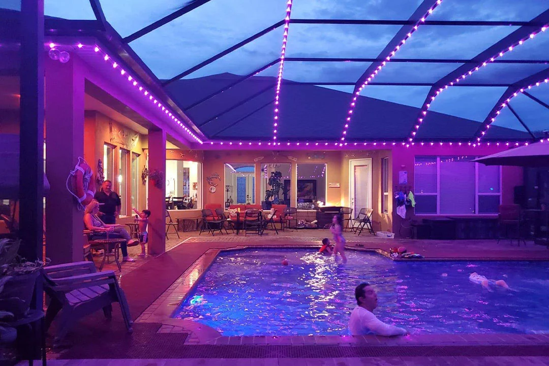 Attached image file People relax and swim in a backyard pool at dusk, surrounded by purple LED lights. Some are sitting by the poolside while others swim, and the enclosed patio area glows warmly through the windows.