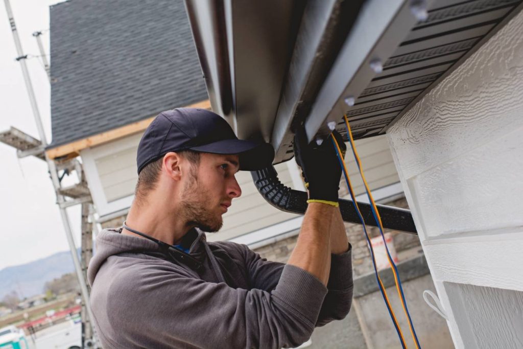 Attached image file A man wearing a cap and gloves installs or repairs a rain gutter on the side of a house, using tools and cords, with a focus on the roofline and exterior siding.