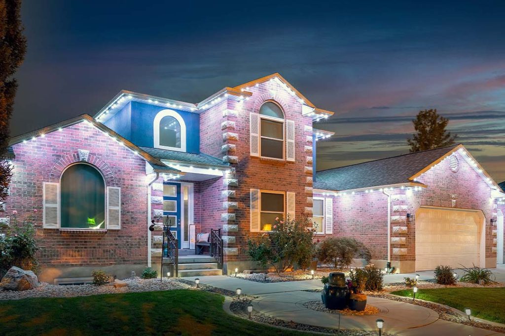 Attached image file Two-story brick house with white trim, illuminated by bright white lights at dusk. The yard has manicured bushes, a curved walkway, and a lit driveway leading to a double garage. The sky is deep blue with pink clouds.