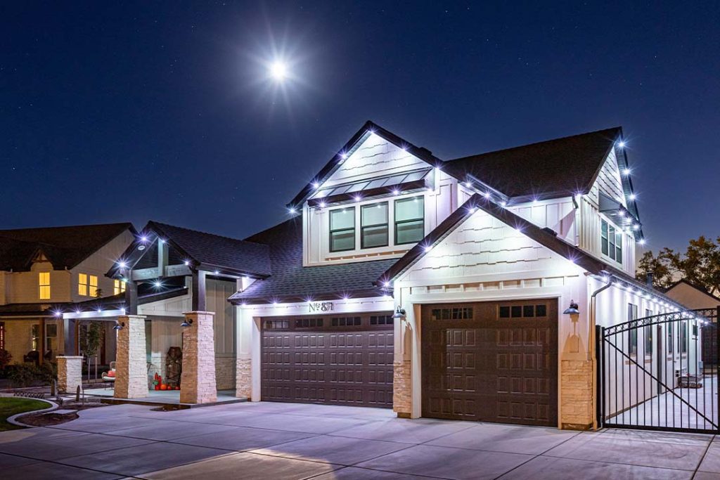 Attached image file A modern two-story house at night, decorated with white string lights along the roofline. The driveway and garage doors are visible, and a bright full moon shines overhead in the clear sky.