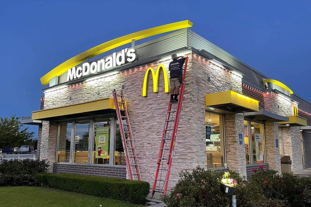Attached image file A McDonalds restaurant at dusk with two ladders propped against the building. A worker is on one ladder, adjusting lights above the entrance. The signature yellow arches and McDonalds sign are clearly visible.