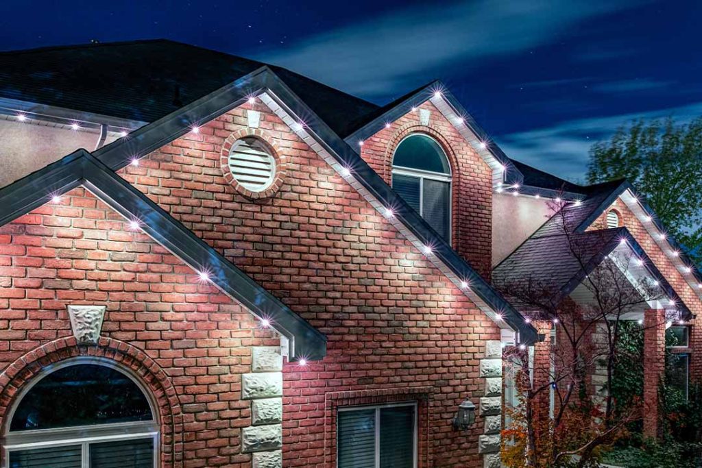 Attached image file A brick house at night with white LED lights outlining its roof and gables, illuminating the exterior against a dark blue sky.