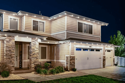 Two-story suburban house at night with stone accents, beige siding, a double garage, and warm outdoor lighting outlining the roof. Small shrubs and plants line the front yard.