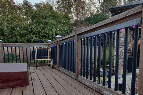 A wooden deck with a railing lined with colorful string lights, a foldable chair, and a brown cushion. Trees and bushes fill the background, and a stone patio is visible beyond the railing.