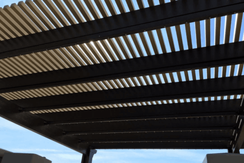 A close-up view from below of a pergola with evenly spaced, dark wooden slats casting striped shadows, with a blue sky and a few scattered clouds visible through the gaps.