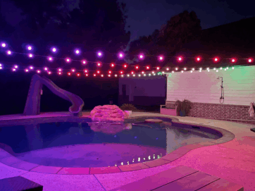 A backyard pool at night is lit by strings of purple, red, and white lights overhead. A slide leads into the pool, and a small rock waterfall feature sits beside it. The area is quiet and empty.