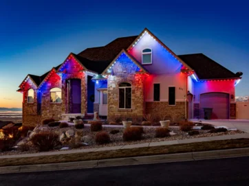 A modern house with stone and brick exterior is decorated with red, white, and blue lights along the roofline at dusk, with a clear evening sky and distant mountains in the background.