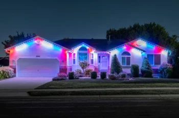 Single-story house at night with bushes in front, illuminated by bright red, white, and blue lights along the roof, windows, and entrance, creating a festive appearance. The driveway and lawn are visible under the lights.