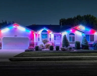 Single-story house at night with bushes in front, illuminated by bright red, white, and blue lights along the roof, windows, and entrance, creating a festive appearance. The driveway and lawn are visible under the lights.
