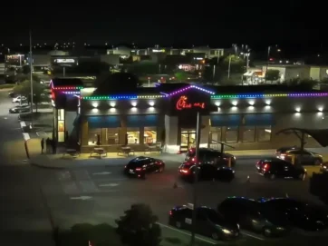 A Chick-fil-A restaurant at night is outlined with colorful lights. Cars line up in the drive-thru, and the parking lot is partially filled. City lights are visible in the background.