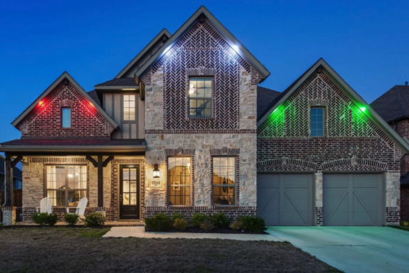 Attached image file A two-story brick and stone house with porch lights, a double garage, and colored lights (red, blue, green) illuminating the exterior, pictured in the evening with a dark blue sky.