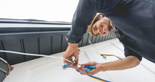 Attached image file A person smiling while installing or repairing electrical wiring on an exterior wall, holding colorful wires and using tools, viewed from a low angle.