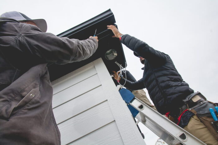 Two people on ladders install wiring under the roof of a small white building. One person wears a jacket and tool belt; both are using tools and working together on a cloudy day.