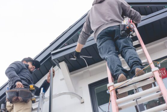 Attached image file Two workers on ladders install wiring under the eaves of a house. One holds tools and wiring, while the other stands on a ladder, reaching up to the roof. Both appear to be wearing tool belts and work clothing.