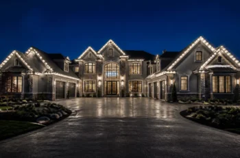 A large, elegant house at night with peaked roofs, stone accents, and bright white lights outlining the roof. The driveway is wide and shiny, reflecting the house’s lights. Manicured bushes and grass surround the home.