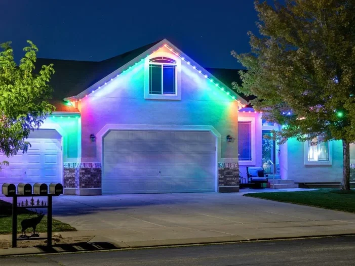 A suburban house at night is decorated with bright, colorful LED lights outlining the roof and garage, illuminating the driveway and trees. Four mailboxes are visible in front.