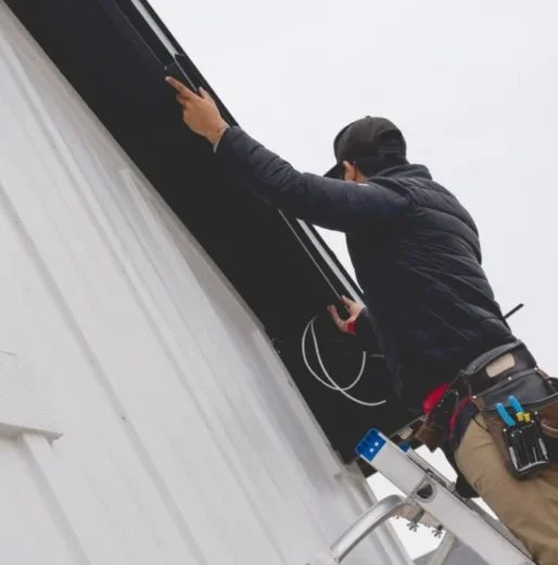 Attached image file A person standing on a ladder installs equipment or wiring near the roof of a white building, wearing a tool belt, jacket, and cap. The sky is overcast.