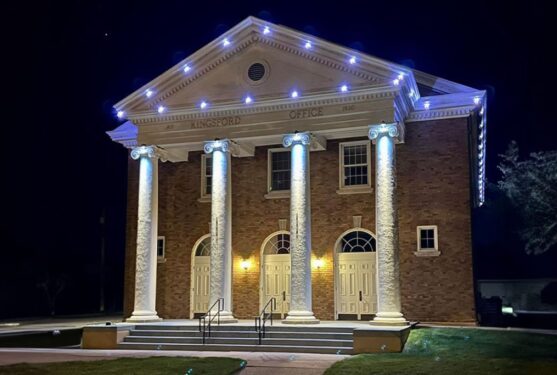 Attached image file A well-lit, two-story brick building with four large white columns and three arched doorways at night. White lights outline the roof, and Kingsford Office is engraved above the columns.