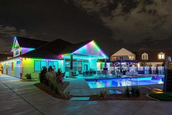 Attached image file A modern house at night with multicolored LED lights outlining the roof, a well-lit swimming pool, lounge chairs, and a landscaped backyard under a partly cloudy sky.