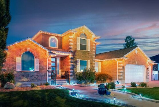 Attached image file A two-story brick house is decorated with string lights along the roofline and illuminated by garden lights at dusk. The yard features a manicured lawn, shrubs, rocks, and a curved walkway leading to the front entrance.