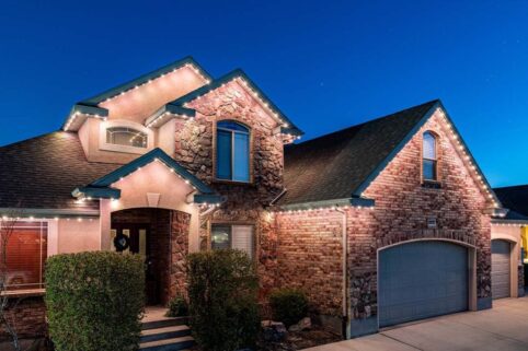 A two-story brick and stone house at dusk, with lit white string lights along the roofline, large windows, bushes near the entryway, and a gray garage door.