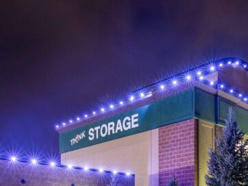 A building at night with THINK STORAGE written on the side, outlined with bright blue lights along the roof. Evergreen trees are visible in the foreground under a cloudy, purple sky.