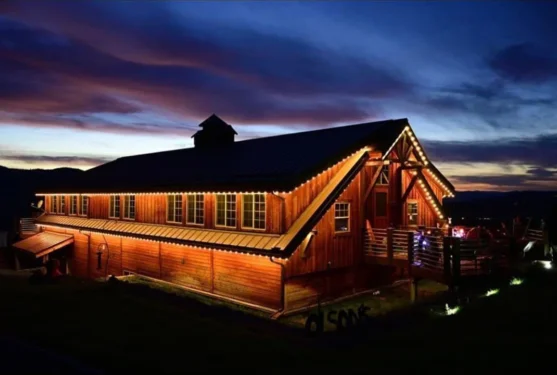 Attached image file A large wooden barn illuminated by warm string lights at dusk, with a dramatic blue and purple sky in the background. The building stands out against the dark landscape.
