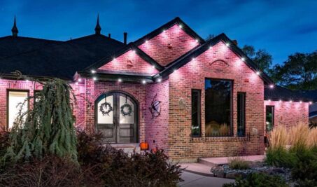 A brick house at dusk with glowing pink and white uplighting along the roofline, arched double doors, two wreaths, tall grasses, shrubs, and a small pumpkin on the porch. The sky is deep blue.