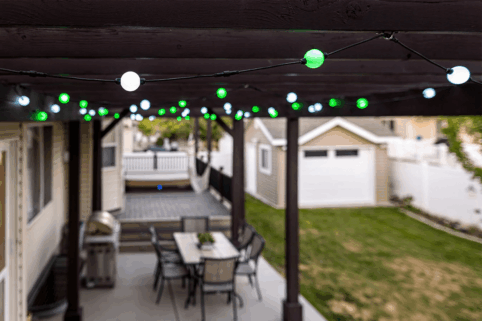 String lights with green and white bulbs hang from a wooden patio pergola above an outdoor dining table and chairs. In the background, there is a backyard with grass and a detached garage.